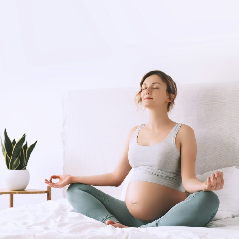 Schwangere Frau meditiert in einer ruhigen Umgebung mit Pflanzen im Hintergrund auf dem Bett.