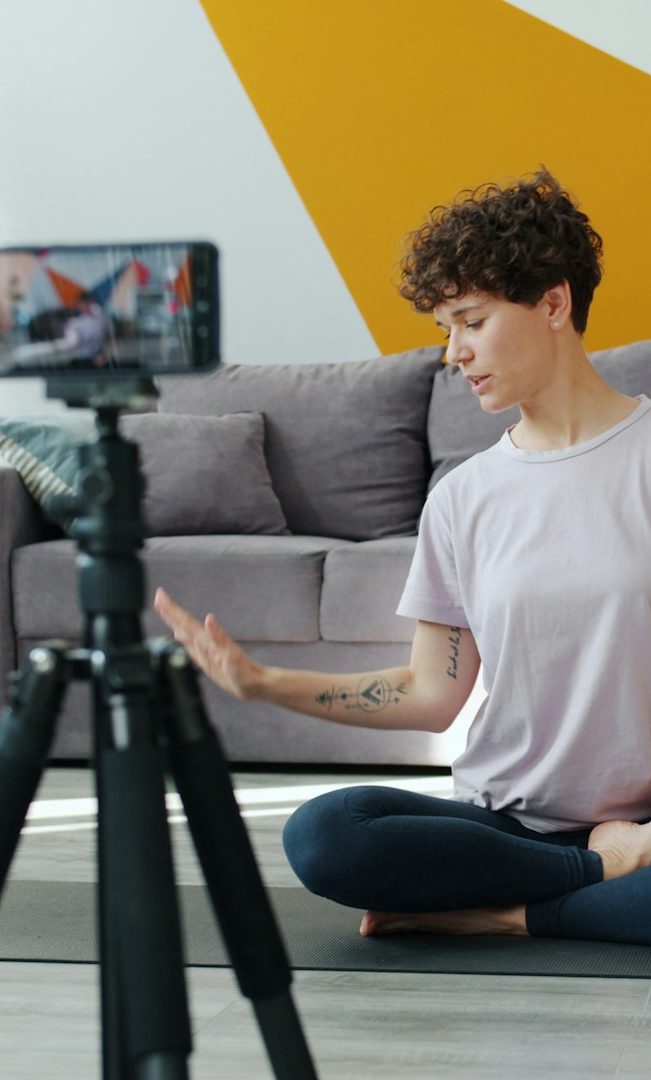 Frau mit lockigem Haar sitzt im Schneidersitz und filmt sich mit einem Smartphone auf einem Stativ.
