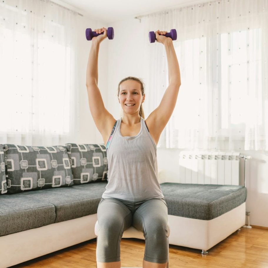Frau mit Hanteln, die auf einem Stuhl im Sitzen Krafttraining macht, in einem hellen Raum.