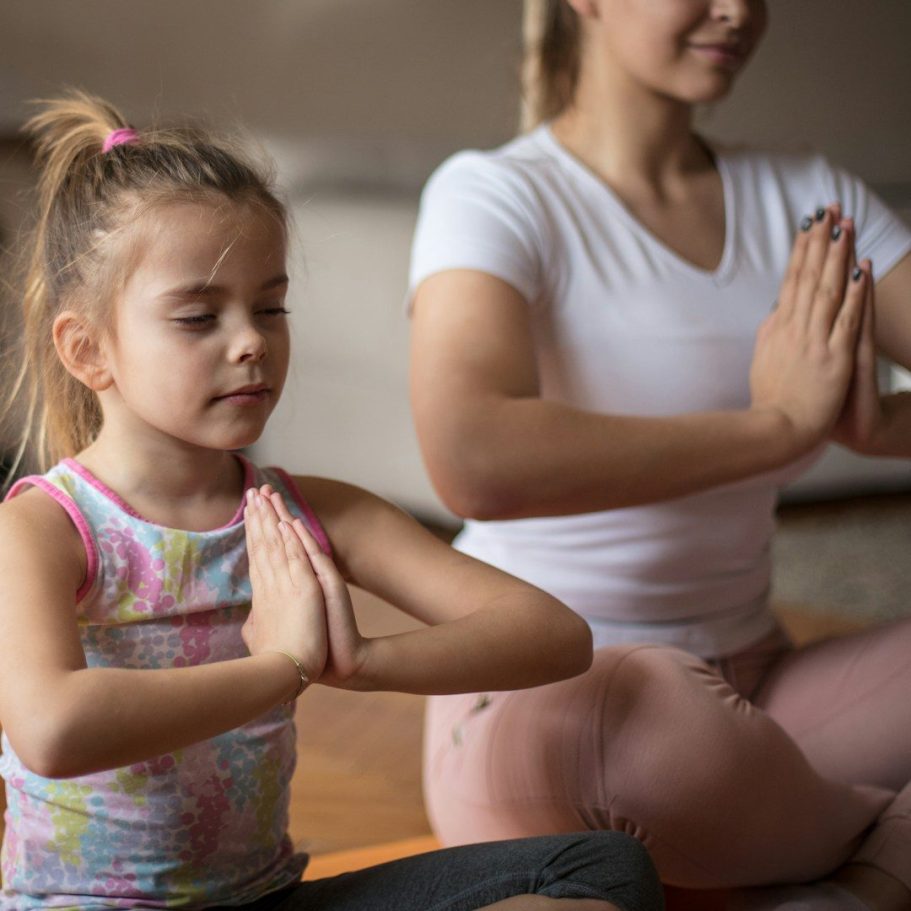 Kind mit seiner Mutter im Schneidersitz machen Yoga und haben die Hände geschlossen zum Herzen zum Namaste