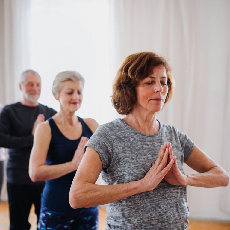 Drei Personen praktizieren Yoga in einem hellen Raum, zwei stehen im Hintergrund.