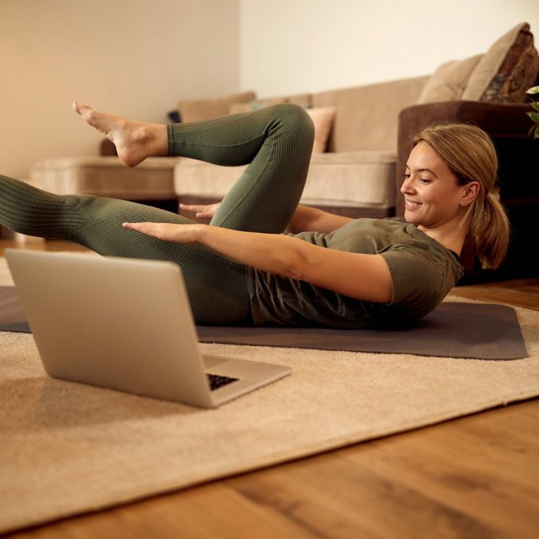 Frau in Sportkleidung macht Übungen auf einer Matte vor einem Laptop.