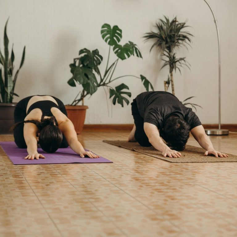 Zwei Personen machen Yoga in einer ruhigen Umgebung mit Pflanzen im Hintergrund.