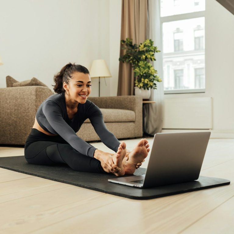 Eine Frau dehnt sich auf einer Matte vor einem Laptop in einem hellen Raum.