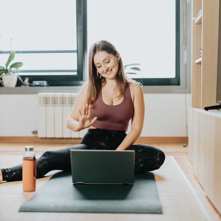 Junge Frau sitzt auf einer Yogamatte, lächelt und winkt vor einem Laptop.