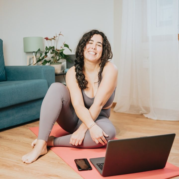 Lächelnde Frau in Sportkleidung sitzt auf einer Yogamatte neben einem Laptop und Smartphone.