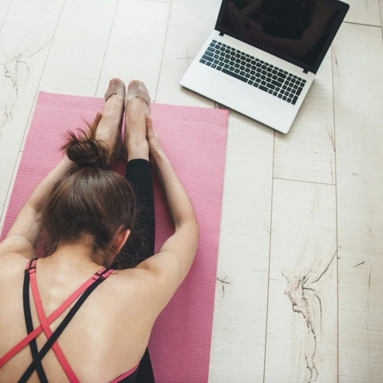 Person macht eine Dehnübung auf einer Matte, ein Laptop steht daneben.